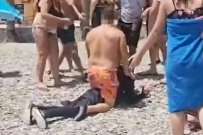 Bañista con bañador naranja inmovilizando en el suelo a una persona en una playa de piedras, rodeado de gente